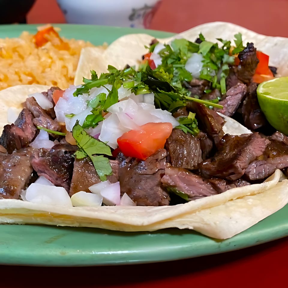 Carne asada tacos with diced onions, cilantro, and a lime wedge, served with rice.