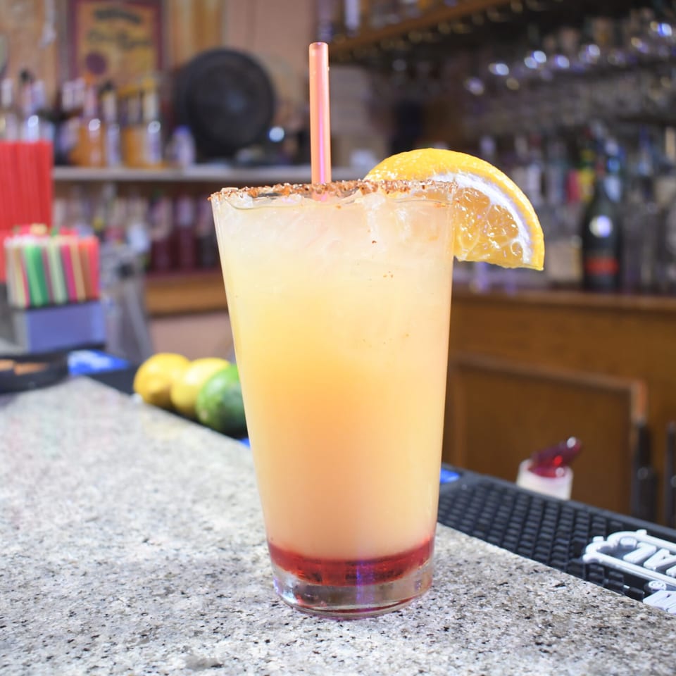 A tall glass of a yellow and red layered drink with a salted rim and orange slice.