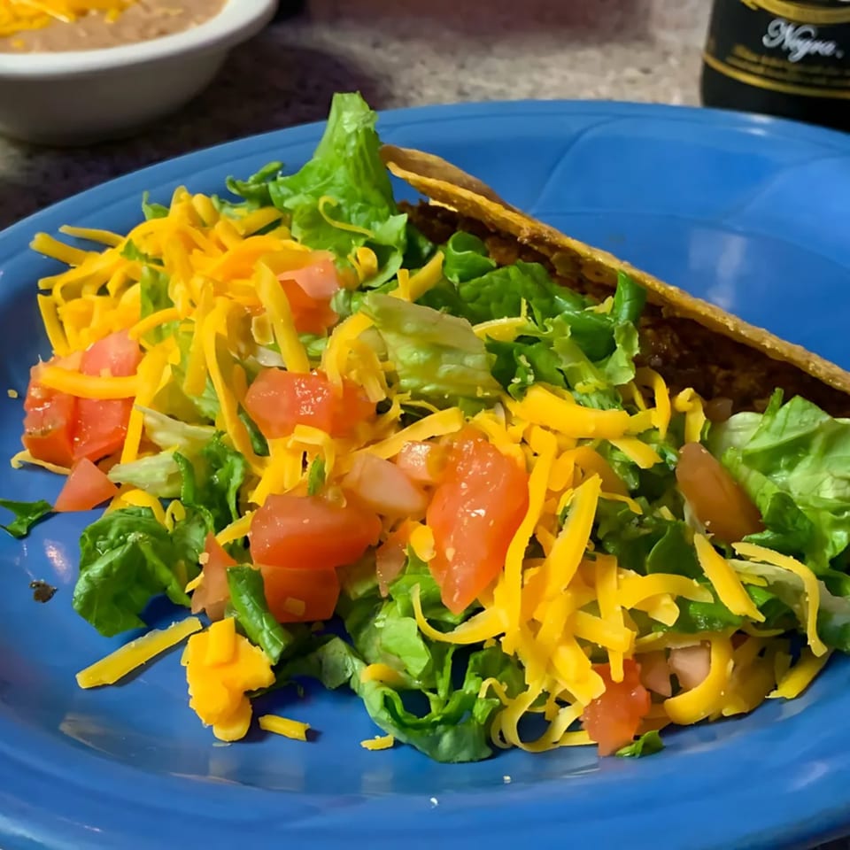 A crispy taco shell filled with ground beef, topped with lettuce, shredded cheese, and diced tomatoes on a blue plate.