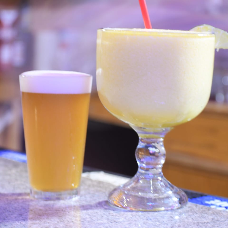 A frosty margarita with a red straw and a beer on a bar counter.