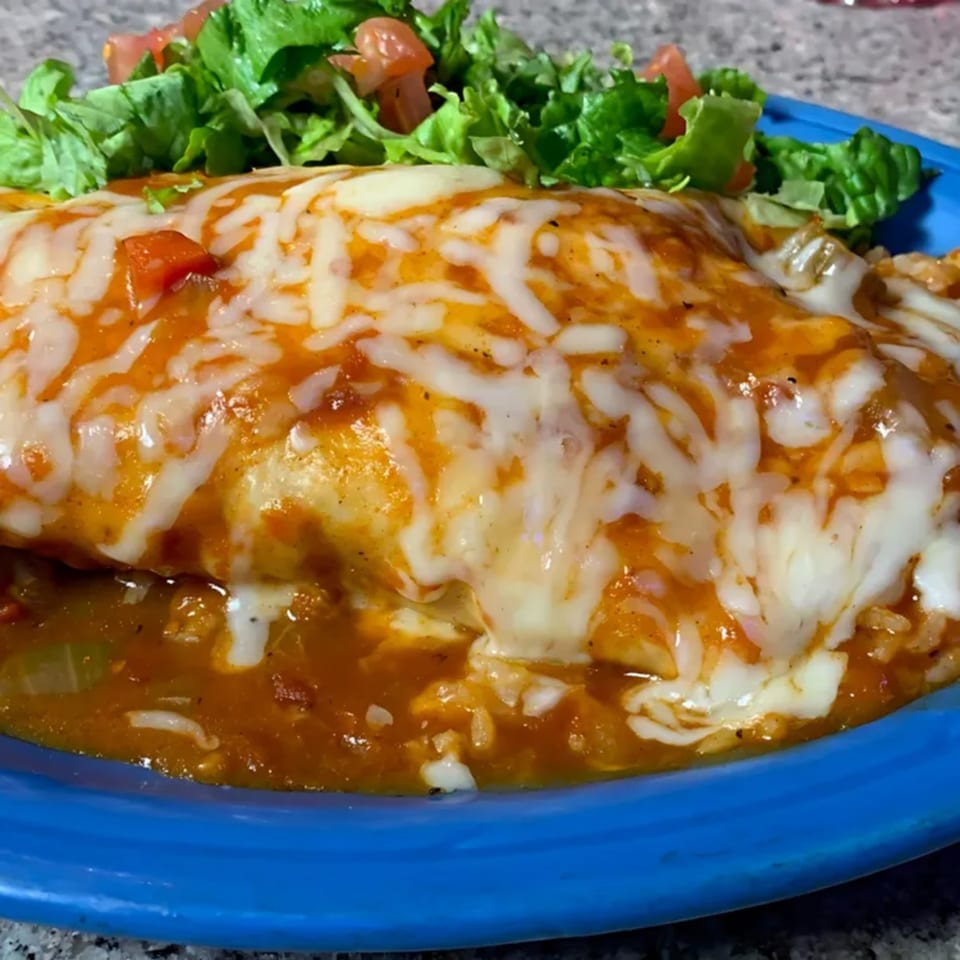 A large, cheesy burrito smothered in red sauce, served with a side of fresh lettuce and diced tomatoes.