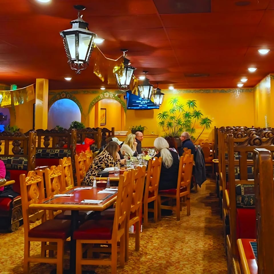 Diners seated at wooden tables with red chairs in a vibrant Mexican restaurant with murals and lanterns.