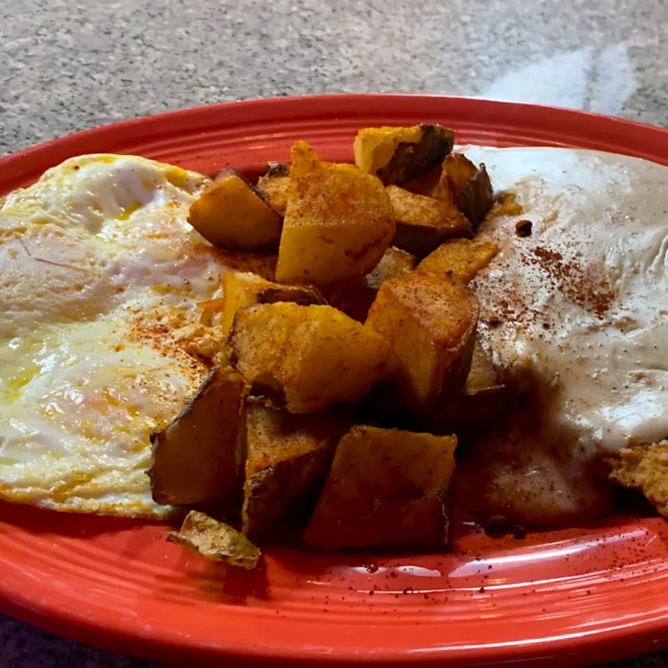 A red plate holds two fried eggs, seasoned potato wedges, and a creamy white sauce.