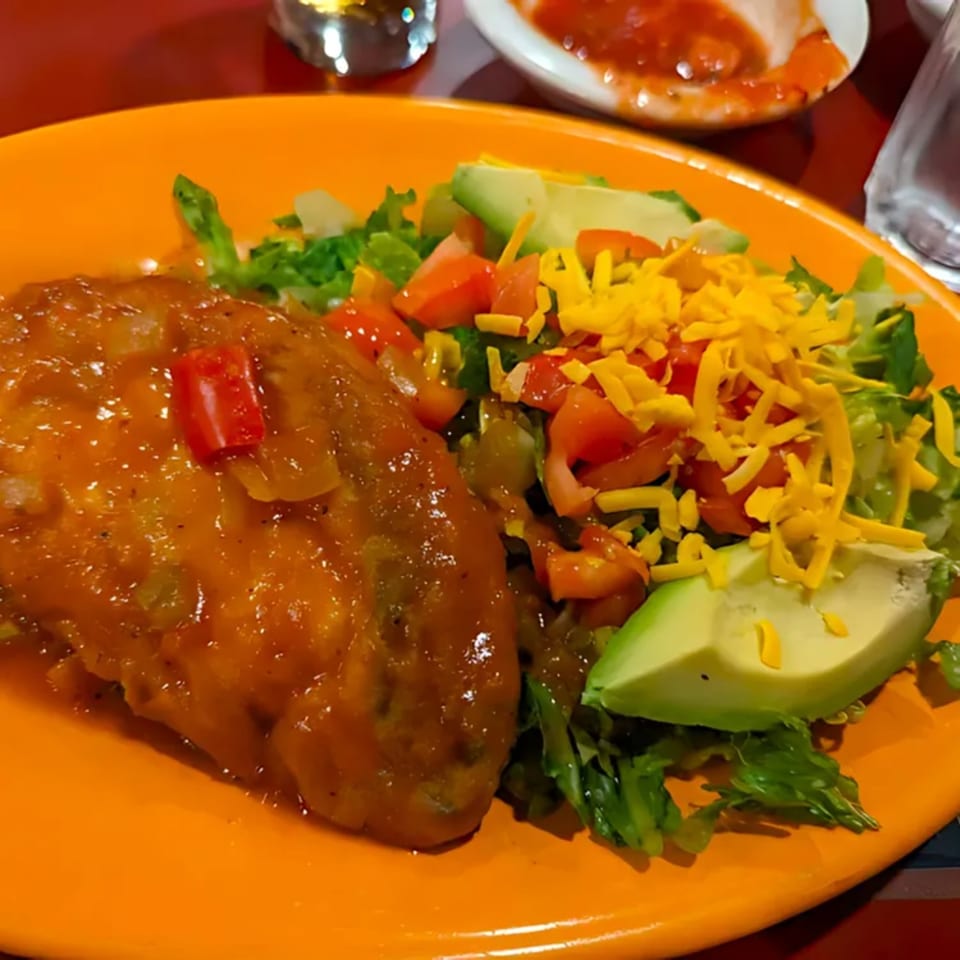 A Mexican dish with a chile relleno, side salad, avocado, and shredded cheese on an orange plate.