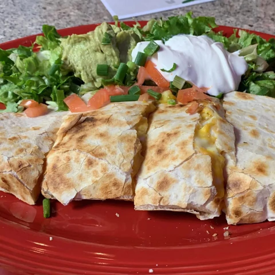 A cheese quesadilla cut into triangles, served with guacamole, sour cream, lettuce, and diced tomatoes.