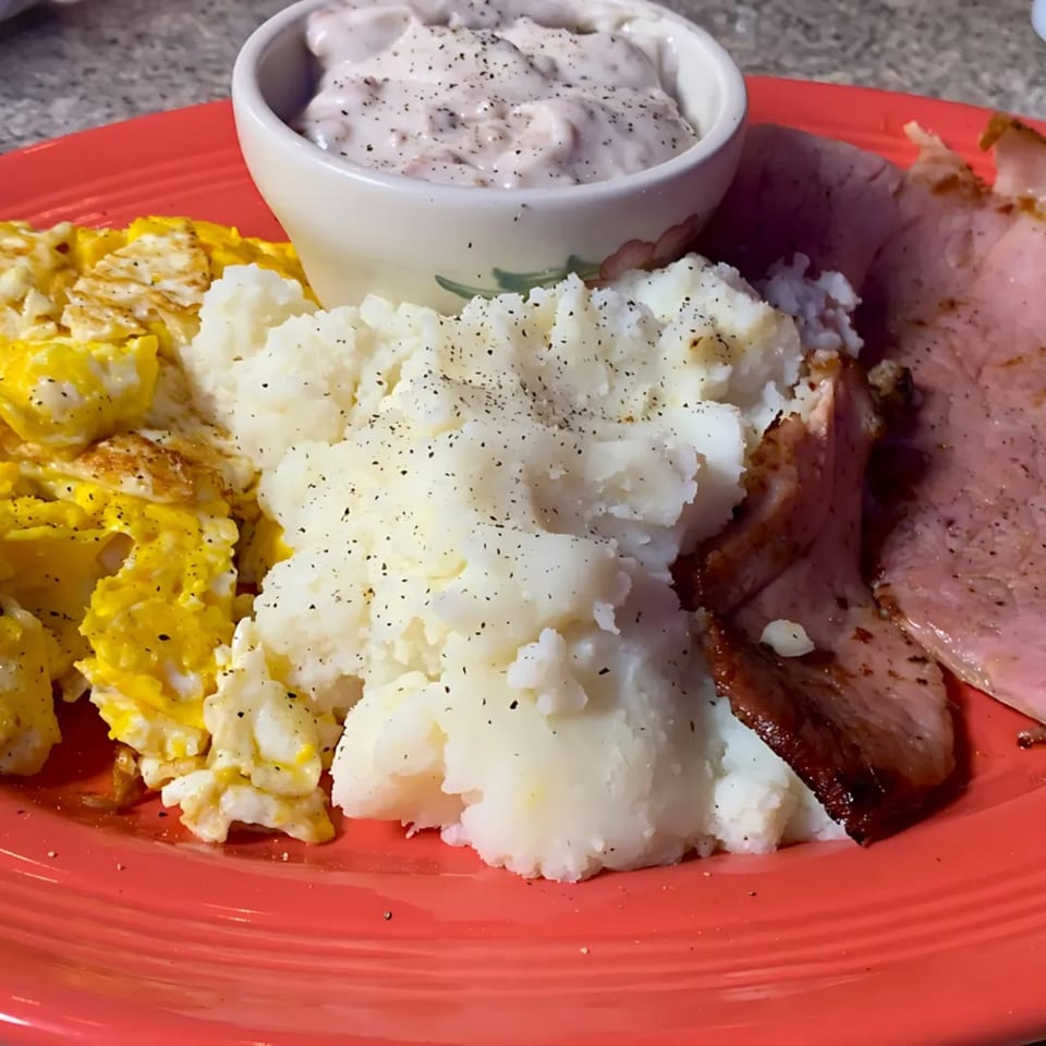 A red plate holds scrambled eggs, mashed potatoes, ham, and a side of creamy gravy.