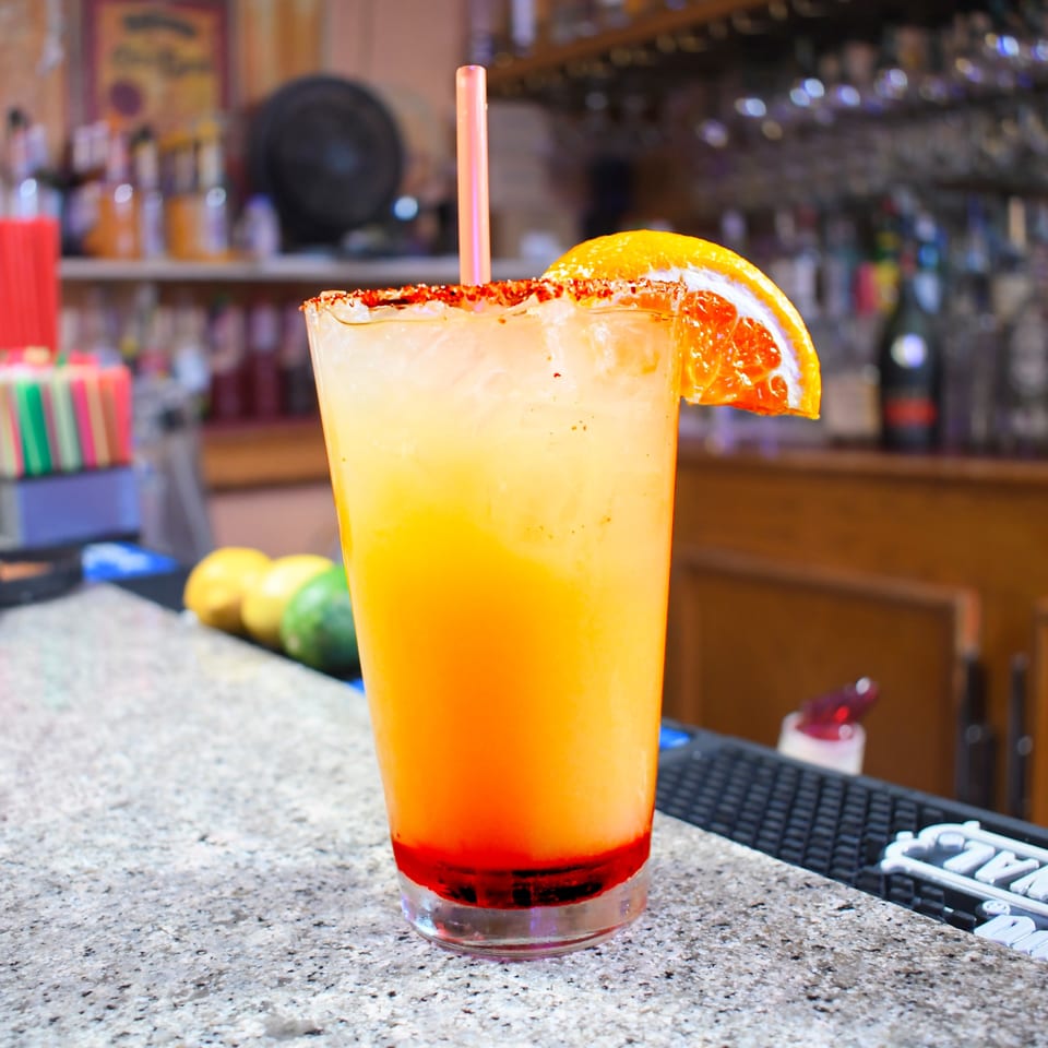 A vibrant orange and yellow drink with a chili-lime rim and orange slice sits on a bar.