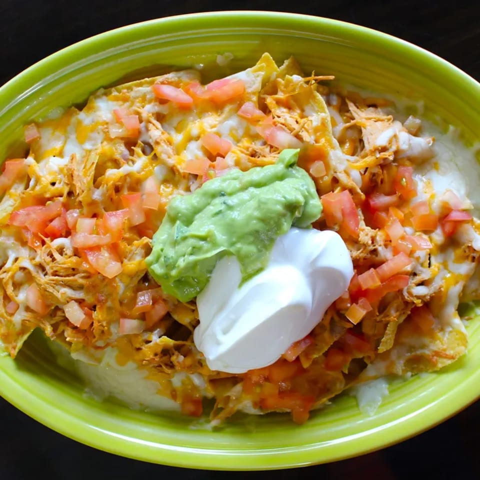 Chicken nachos topped with melted cheese, diced tomatoes, guacamole, and sour cream in a green bowl.